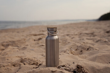 Steel thermos water bottle standing in the sand on the beach on a beautiful summer day at the coast...