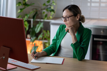 Focused female freelancer working on future business project, makes plan writing in notebook. Concentrated woman in glasses looking at computer monitor learns details in cozy home office with plants. 