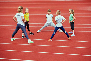 Fototapeta premium Female coach training athletes. Group of children running on treadmill at the stadium. Concept of sport, achievements, studying, goals, skills. Little boys and girls training outdoor.