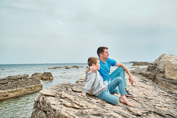 Fototapeta na wymiar Happy family, father and son bonding, sitting on stone by the sea looking at view enjoying summer vacation. Togetherness Friendly concept