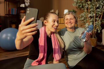 Happy woen laughing looking at cell phone talking with father by video link wearing sportswear relaxing after hard yoga lesson online indoors. Hapy eldelry woman having fun with young daughter.