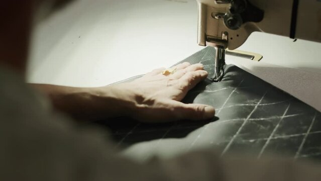Unrecognizable factory worker making black leather car seat covers on sewing machine in workshop