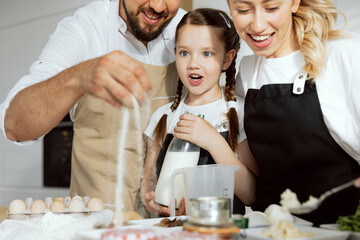Close-up shot surprised daughter with braids holding milk in hands looks at how father sprinkling flour into bowl. preparing pizza ingredients. Delighted mom holding spoon with food. Family baking