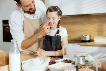 Curious daughter and father in aprons spending time together cooking baking in modern kitchen. Happy father touching daughter's nose. Pizza ingredients on wooden surface salami flour milk eggs butter.