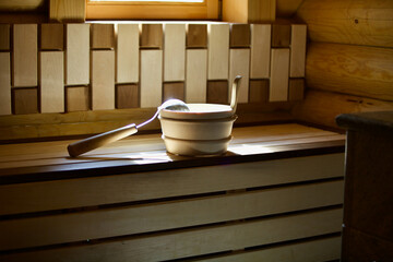 Cozy wooden interior of wood burning sauna and sauna accessories close-up.  A wooden bucket, felt mitten, ladle and bamboo broom on a wooden bench. Closeup of sauna accessories in the sunshine. SPA.