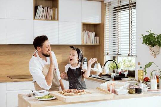 After Dressing Homemade Pizza Father Giving High Five Delighted Kid Wearing Apron. Happy Parent Teaching Daughter Baking Cooking Homemade Pizza. Pizza Ingredients On Wooden Surface.