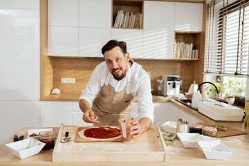 Handsome young father husband chef applying tomato sauce on homemade pizza dough looking at camera smiling while preparing. Man preparing birthday dinner for happy chidren kids.