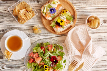 Aesthetic pescetarian table - toasts with salmon, cottage cheese, edible flowers. Mediterranean salad with fresh vegetables and salmon. Cup of tea. Atmospheric autumn lunch time