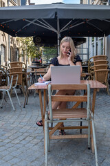 Attractive young blonde business woman using her laptop while calling with her smartphone, sitting outdoors in a European city at the typical Belgian cafe terrace. High quality photo