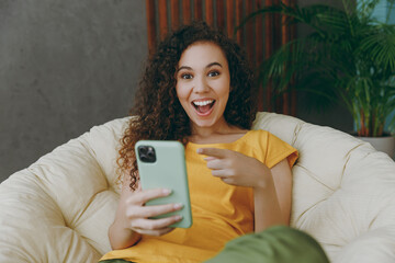 Young woman of African American ethnicity in casual clothes use point finger on mobile cell phone sits in armchair stay at home flat rest relax spend free spare time in living room indoors grey wall