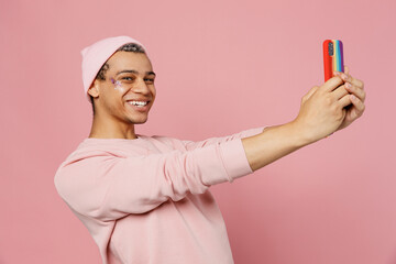 Side view young gay man wear sweatshirt hat do selfie shot on mobile cell phone post photo on social network isolated on plain pastel light pink color background studio Lifestyle lgbtq pride concept.