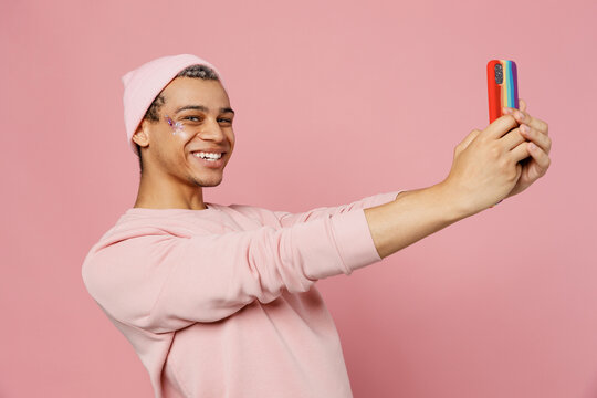 Side view young gay man wear sweatshirt hat do selfie shot on mobile cell phone post photo on social network isolated on plain pastel light pink color background studio Lifestyle lgbtq pride concept.