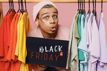 Young fun gay man wear sweatshirt hat hold card sign with black friday title text near rack with t-shirts at store isolated on plain pastel light pink background studio Lifestyle lgbtq pride concept