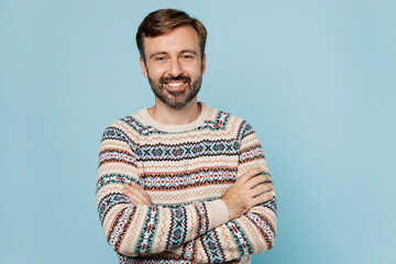 Young cheerful fun happy smiling caucasian man 30s he wear sweater hold hands crossed folded look camera isolated on plain pastel light blue cyan background studio portrait. People lifestyle concept.