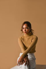 Obraz premium Fashion studio photo, Autumn and Winter concept. Attractive young woman in trendy sweater sitting over brown studio background