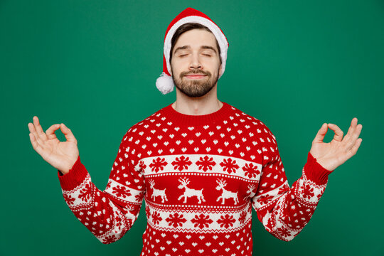 Merry young man 20s wear red warm knitted sweater Santa hat posing spread  hands in yoga om aum gesture relax meditate isolated on plain dark green  background New Year 2023 holiday celebration