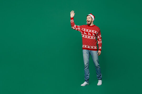 Full body smiling happy fun merry young man wear red warm knitted sweater Santa hat posing waving hand say hi isolated on plain dark green background studio. New Year 2023 holiday celebration concept.