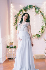 Fototapeta premium Young pregnant woman with dark hair in airy dress in a room decorated with pine needles and sparkling garlands for Christmas. Christmas mood . Pregnancy
