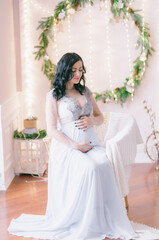 Fototapeta na wymiar Young pregnant woman with dark hair in airy dress in a room decorated with pine needles and sparkling garlands for Christmas. Christmas mood . Pregnancy