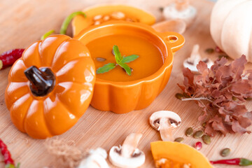 Warm and tasty pumpkin soup with mint on top in pumpkin shaped bowl with spoon surrounded by green and red peppers, mushrooms, garlics, pumpkin seeds on wooden table. High quality photo