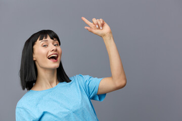 an admiring woman stands on a gray background in a blue T-shirt smiling happily raising her hand...