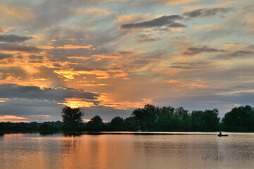 sunset over the lake in the meadow with reflection in the water of the sky with clouds, fisherman, reeds, tree silhouettes