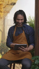 Happy young man entrepreneur wearing apron holding tablet device smiling. Person checking inventory...