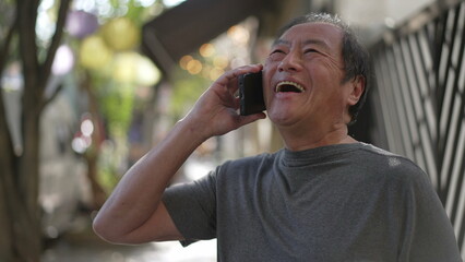 Fototapeta na wymiar Happy senior man receiving great news on phone standing in city street. Authentic real life laugh and smile of an older Asian person speaking with cellphone laughing