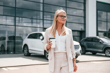 Standing outdoors near new automobile. Woman in white formal clothes is in the car dealership