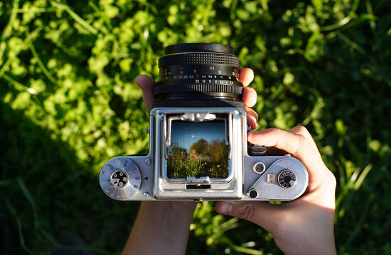 A photographer holding a vintage analog photo camera focusing ajusting taking pictures outside looking through a large square format viewfinder mechanical settings solid metal cogs garden background
