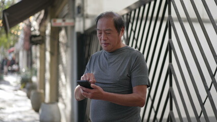Fototapeta na wymiar An Asian senior man using cellphone standing in city street. Older person holding smartphone device browsing internet online in daylight