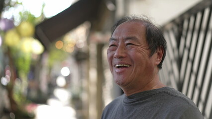 A happy Senior Asian man standing in street sidewalk smiling. Portrait of a middle aged older joyful person in daylight. Authentic real life smile