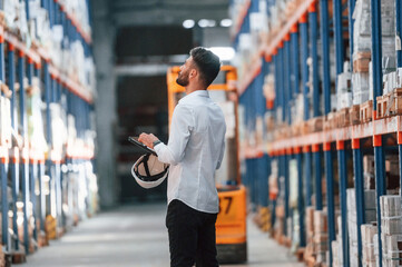 With tablet in hands. Storage worker is in the warehouse with bunch of products