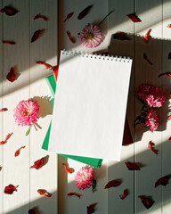 An open blank notepad is on a stack of books on a white wooden background. Nearby are aster flowers and scattered red petals. Floral concept of keeping a diary, making lists, taking notes. Copy space.