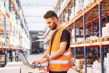 Operating the laptop. Storage worker is in the warehouse with bunch of products
