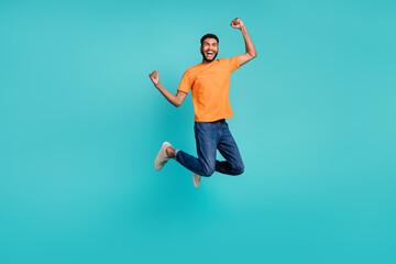 Fototapeta premium Full length photo of funky lucky man dressed orange t-shirt rising fists jumping high isolated turquoise color background