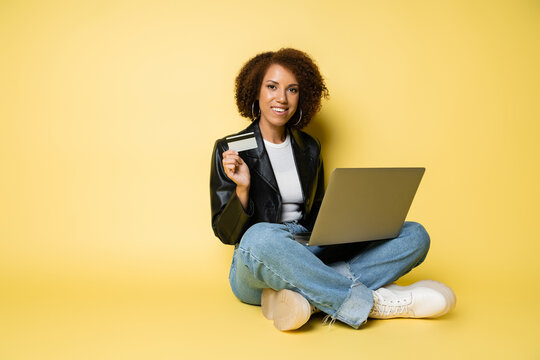 cheerful african american woman in leather jacket holding credit card while sitting with laptop on yellow.