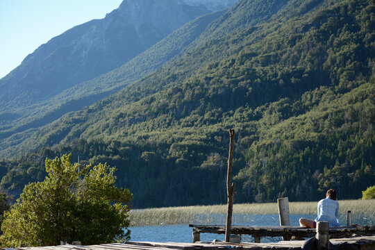 female person sitting on a pier, with a lake and mountains in the background © Lautaro