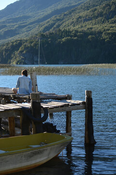 female person sitting on a pier, with a lake and mountains in the background and a boat
