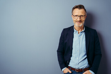 Confident middle-aged man in blue blazer and blue shirt, wearing glasses, standing against plain blue background with copy space and looking at camera - 536062543