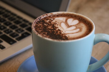 Cup of coffee on a table with laptop in the background. Hot chocolate in a mug. Latte art with a heart. Hot drinks for breakfast at the cafe. Working at the cafe. Laptop and coffee.