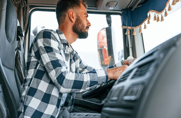 Sitting in the cabin by steering wheel. Young truck driver is with his vehicle at daytime