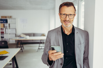 smiling businessman using smart phone standing in office hallway, smiling male professional holding...