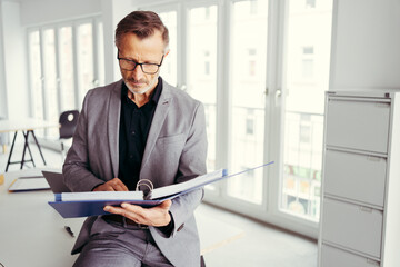older businessman with glasses sitting on desk holds a folder and read the documents - Powered by Adobe