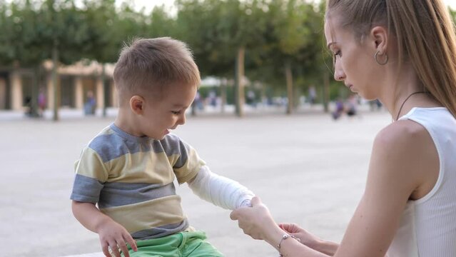 Young mother comforting her little son and giving him bandage in a wounded hand after getting hurt when fall down in street, close-up in the city park. First aid for kids after injury,trauma