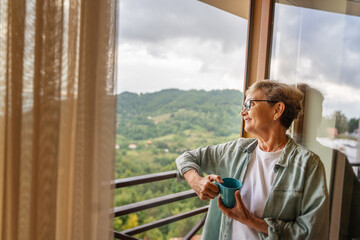 Beautiful happy gray-haired mature senior woman standing at the window at home with a mug of coffee enjoying the scenery