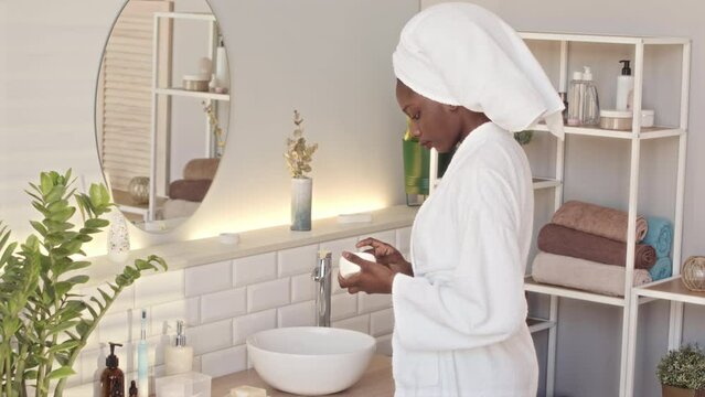 Medium slowmo of young beautiful serene African American woman in white bathrobe and towel on her head standing in front of mirror in luxury bathroom applying moisturizing face cream
