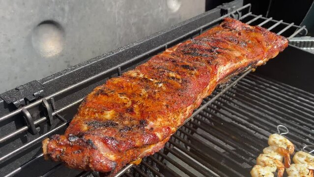 Man on a summer backyard day grilling a rack of babyback ribs and skewered shrimps as he removes one shrip to taste