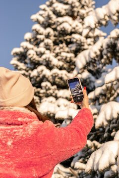 Young woman in pink coat takes pictures of snow-covered fir trees on her smartphone.Winter,technology,social media concept.Selective focus,view behind.