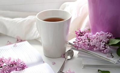The postcard is beautiful. A mug of tea, an old book and a bouquet of purple lilac. A beautiful still life. Spring time. The concept of Good morning.